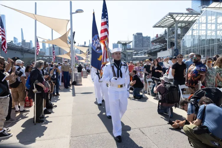Active duty military personnel, matched and holding the American flag, are flanked by crowds on both sides.