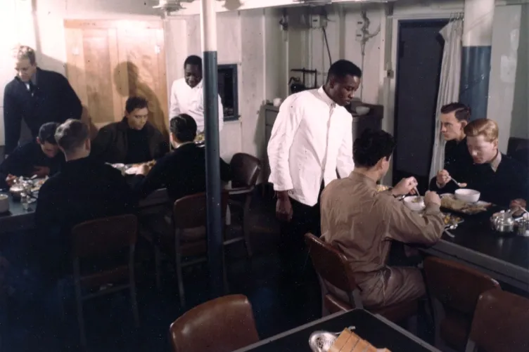 A group of men sitting at a table, enjoying a meal together, while two men stand beside the table, serving food to the group.