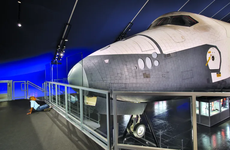 A young boy sits on the floor, looking up at the nose of the space shuttle Enterprise.