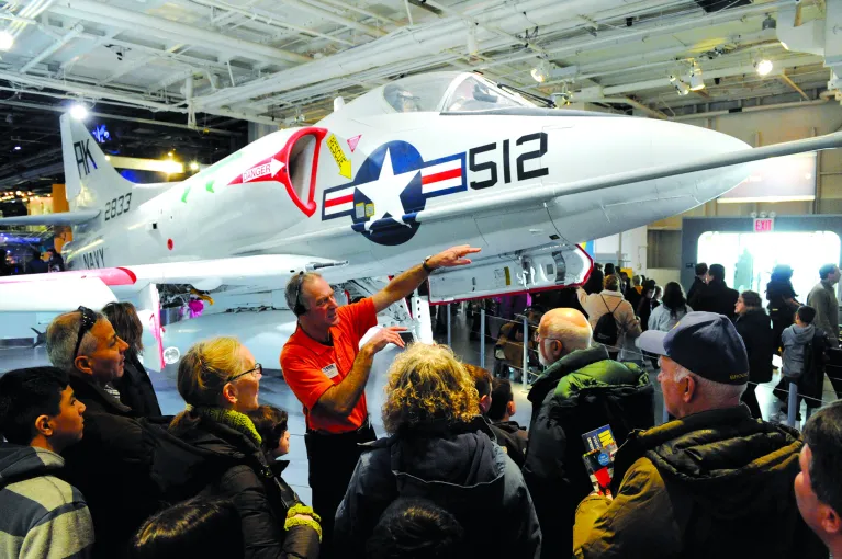 A tour guide is gesturing to a group of visitors in front of an airplane.