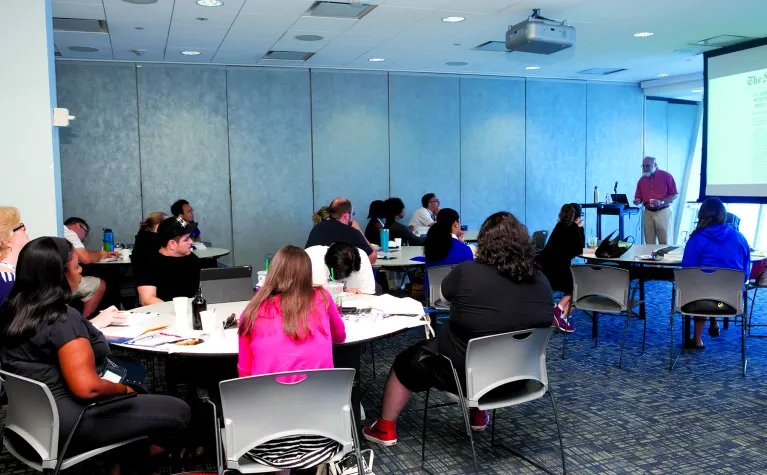 People are seated at round tables in a classroom at the Museum and someone is at a podium giving a presentation