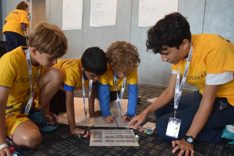Three boys in yellow t-shirts kneel on the ground looking intently at an Ozobot code sheet as a teen on the right points out an example